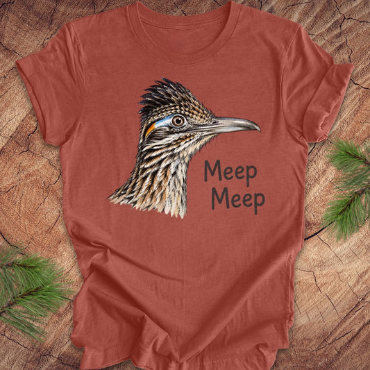 A terracotta-colored short-sleeve t-shirt featuring a detailed graphic of a roadrunner bird and the text 'Meep Meep' printed on the front, laid flat on a rustic wood background with pine branches in the corners.