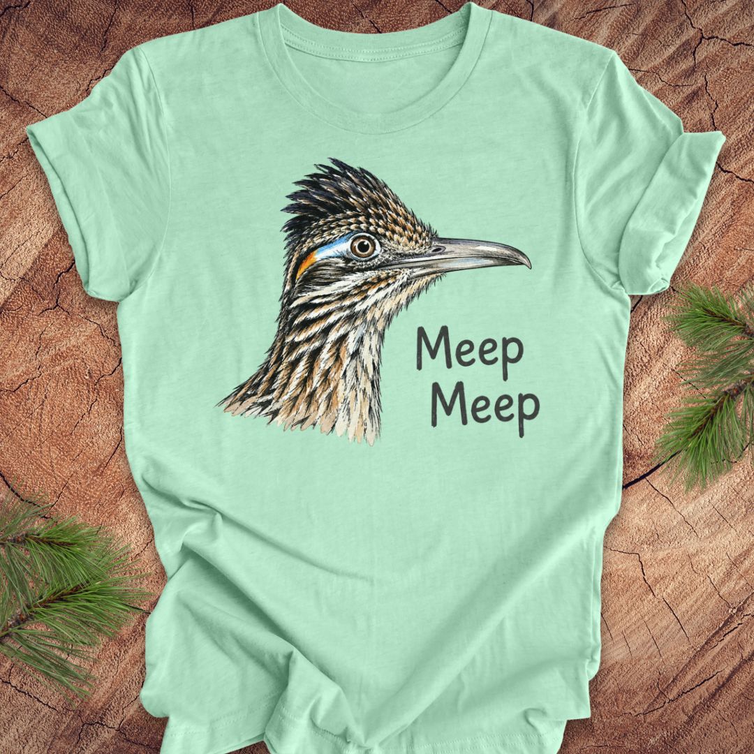 A mint green short-sleeve t-shirt with a detailed graphic of a roadrunner bird and the text 'Meep Meep' printed on the front, laid flat on a rustic wood background with pine branches in the corners.