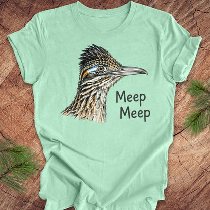 A mint green short-sleeve t-shirt with a detailed graphic of a roadrunner bird and the text 'Meep Meep' printed on the front, laid flat on a rustic wood background with pine branches in the corners.