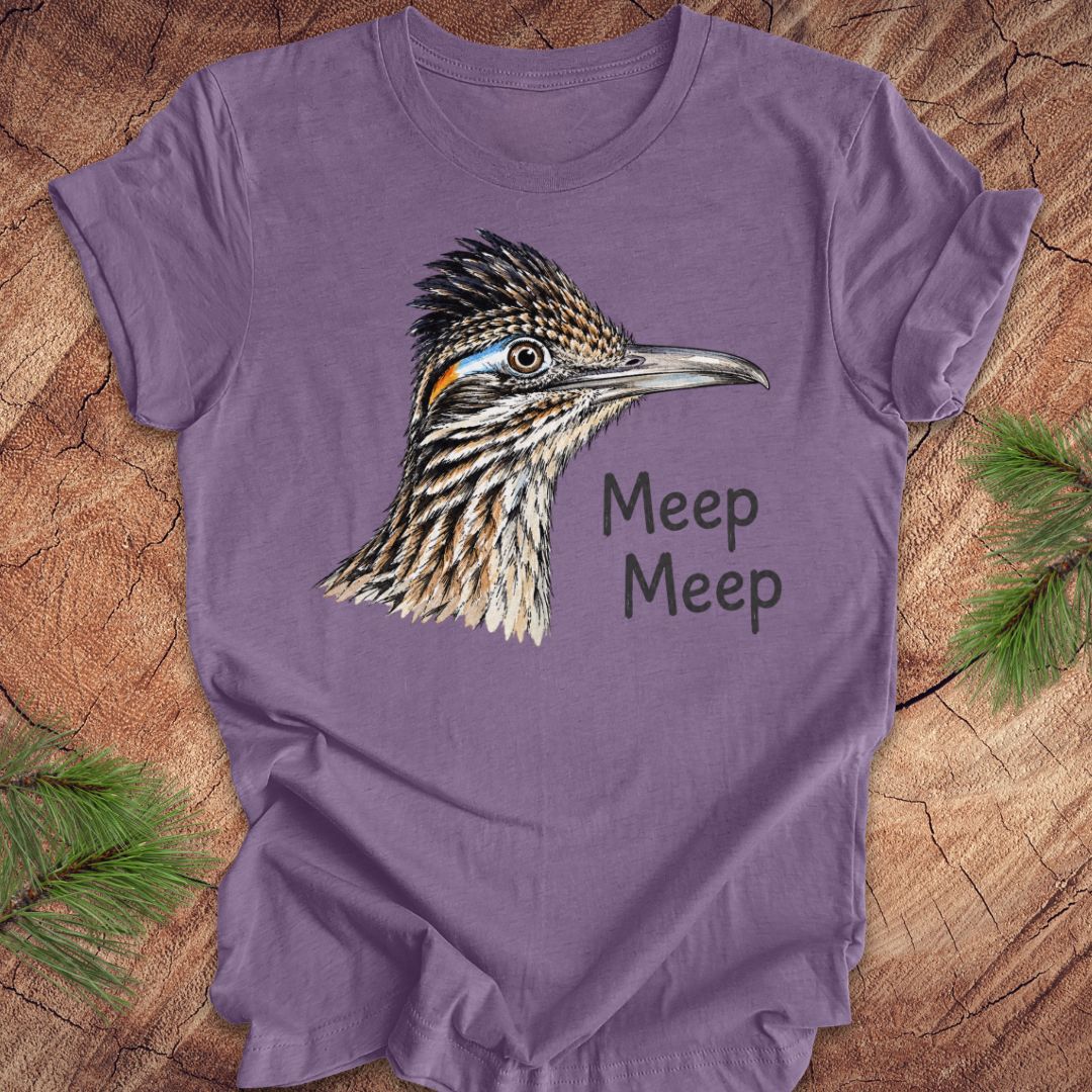 A purple short-sleeve crewneck t-shirt featuring a detailed illustration of a roadrunner bird and the text 'Meep Meep' printed on the front, laid flat on a rustic wood background with pine branches in the corners.