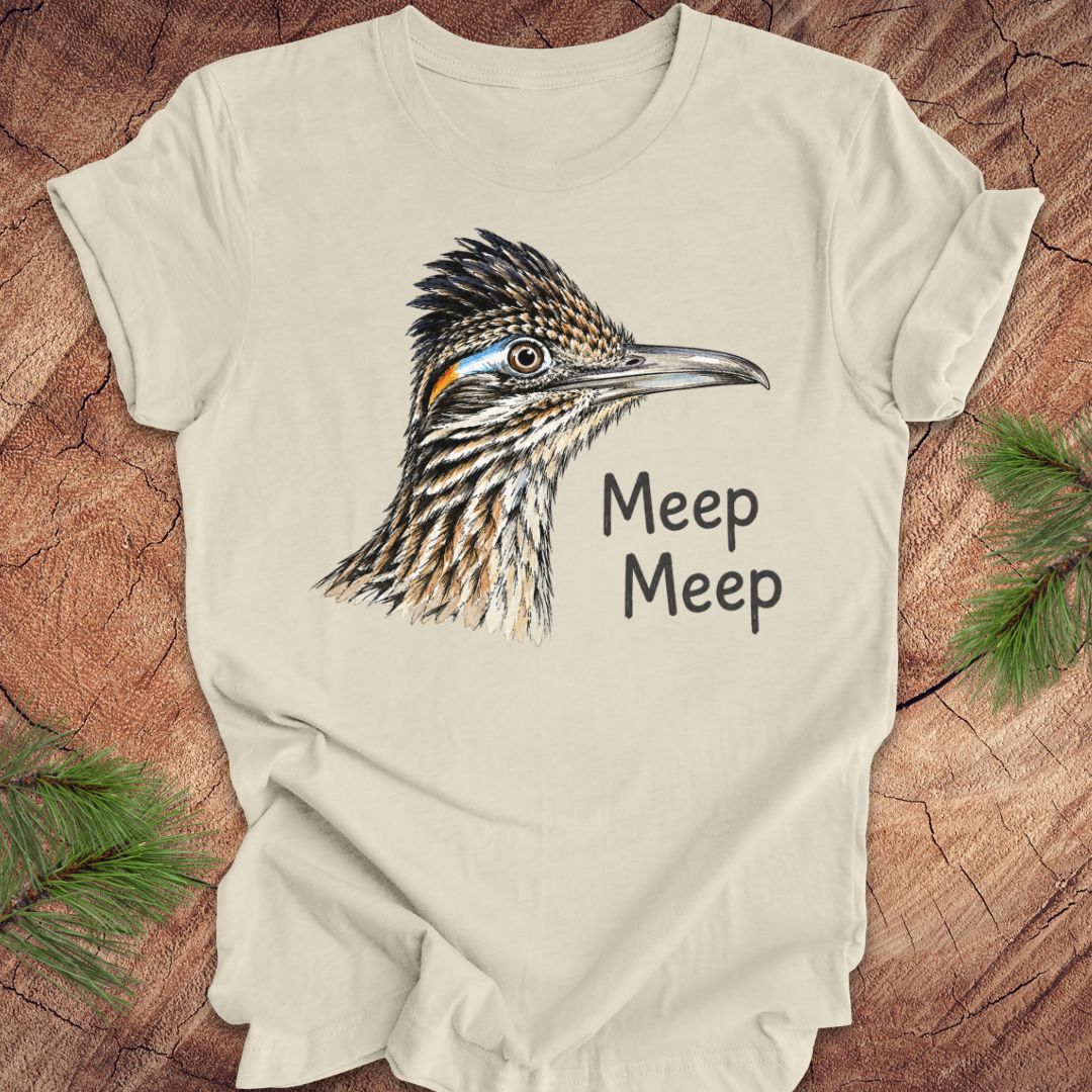 A cream-colored short-sleeve t-shirt featuring a detailed graphic of a roadrunner bird and the text 'Meep Meep' printed on the front. The shirt is laid flat on a rustic wood background with pine branches in the corners.