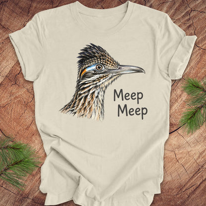 A cream-colored short-sleeve t-shirt featuring a detailed graphic of a roadrunner bird and the text 'Meep Meep' printed on the front. The shirt is laid flat on a rustic wood background with pine branches in the corners.