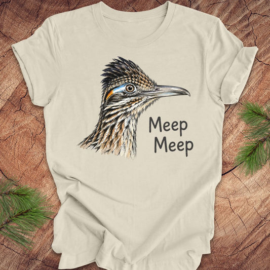 A cream-colored short-sleeve t-shirt featuring a detailed graphic of a roadrunner bird and the text 'Meep Meep' printed on the front. The shirt is laid flat on a rustic wood background with pine branches in the corners.