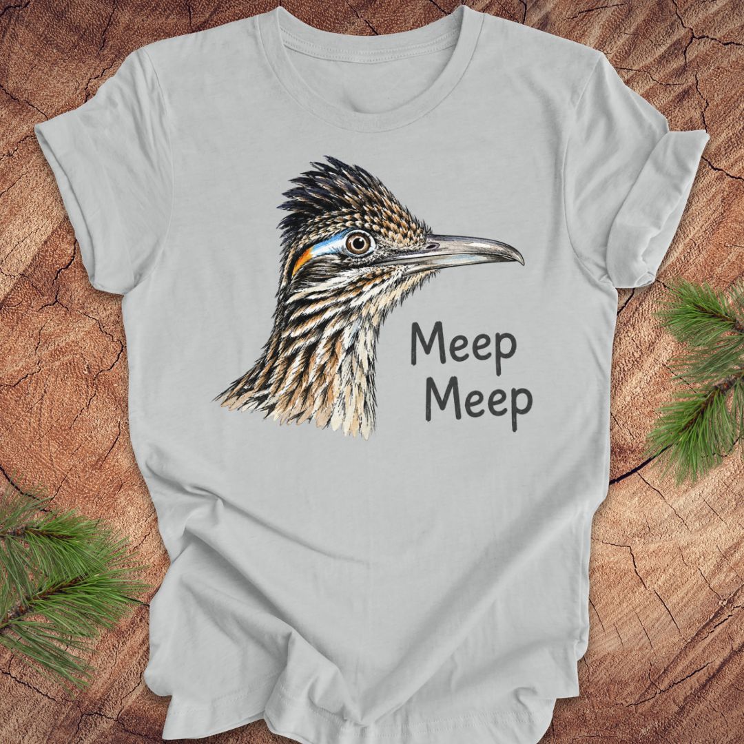 A light gray short-sleeve t-shirt laid flat on a rustic wood background with pine branches in the corners. The shirt features a detailed graphic of a roadrunner bird head with the text 'Meep Meep' printed next to it.