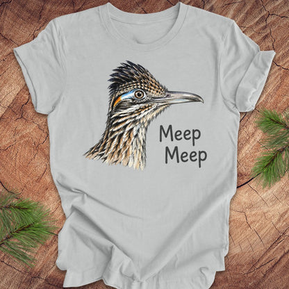 A light gray short-sleeve t-shirt laid flat on a rustic wood background with pine branches in the corners. The shirt features a detailed graphic of a roadrunner bird head with the text 'Meep Meep' printed next to it.