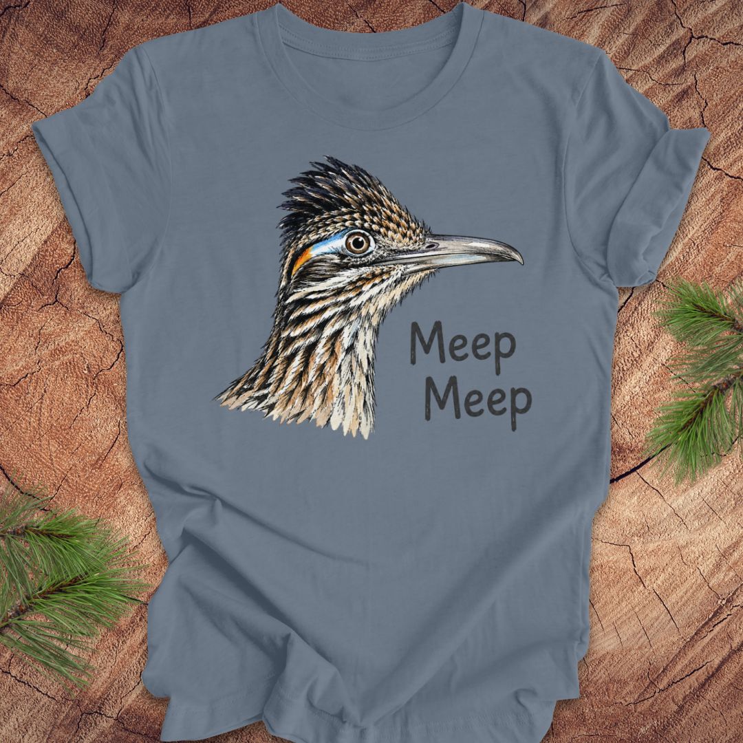 A slate blue crewneck t-shirt featuring a detailed graphic of a roadrunner bird and the text 'Meep Meep' printed on the front, styled flat on a rustic wood background with pine needles.