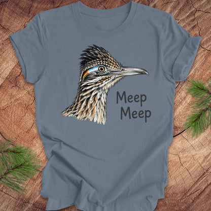 A slate blue crewneck t-shirt featuring a detailed graphic of a roadrunner bird and the text 'Meep Meep' printed on the front, styled flat on a rustic wood background with pine needles.