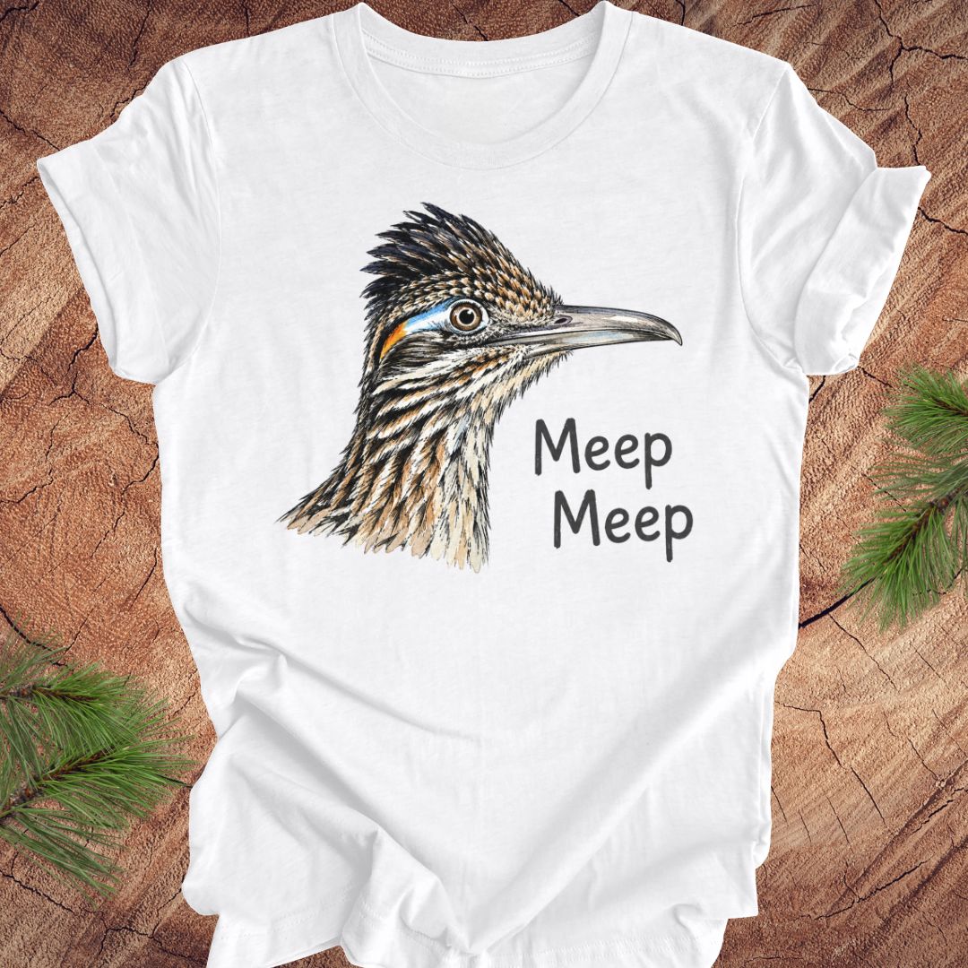 A white short-sleeve crewneck t-shirt featuring a detailed illustration of a roadrunner bird head and the text 'Meep Meep' printed on the front, laid flat on a rustic wood background with pine branches in the corners.