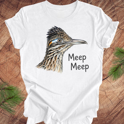 A white short-sleeve crewneck t-shirt featuring a detailed illustration of a roadrunner bird head and the text 'Meep Meep' printed on the front, laid flat on a rustic wood background with pine branches in the corners.