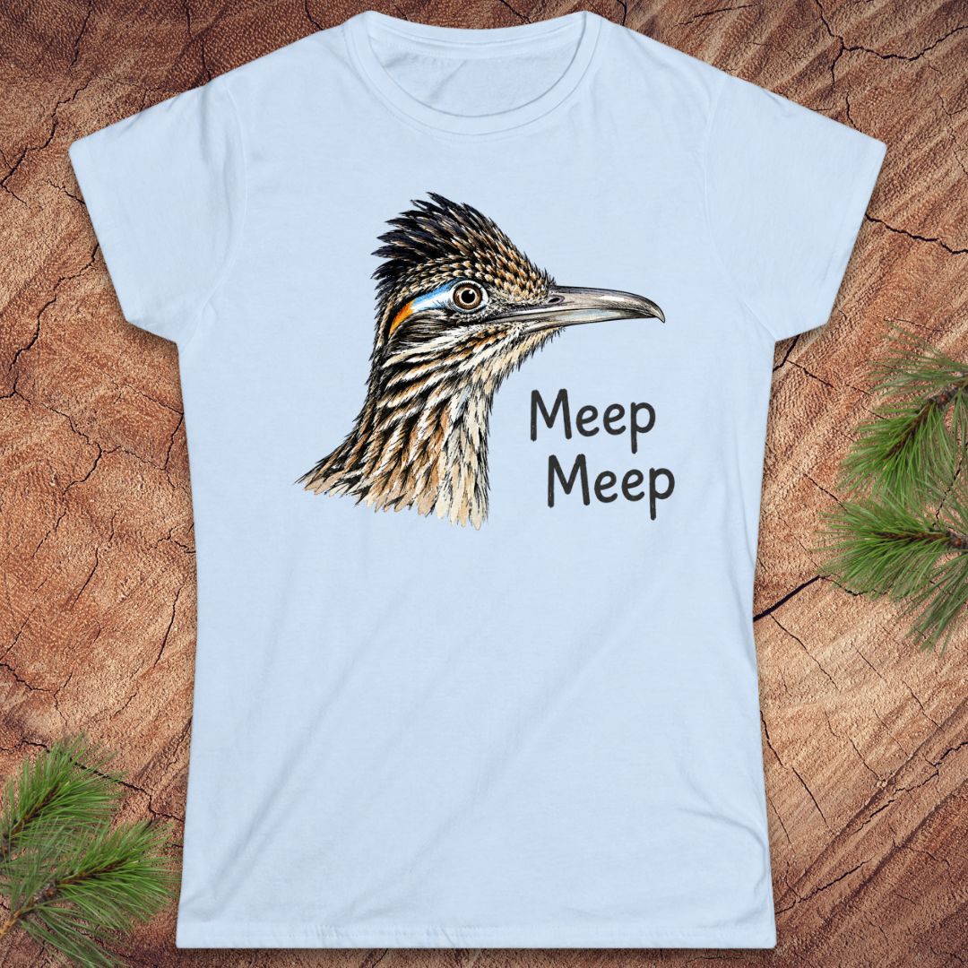 A light blue short-sleeve crewneck t-shirt featuring a detailed graphic of a roadrunner bird head and the text 'Meep Meep' printed in black. The t-shirt is displayed flat on a textured brown wood background with pine needles in the corners.