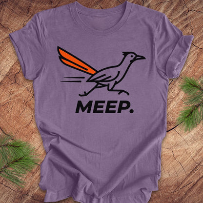 A lavender purple short-sleeve t-shirt laid flat on a rustic wood background with pine needles in the corners. The shirt features a graphic of a black and white roadrunner with an orange tail, above the word 'MEEP.