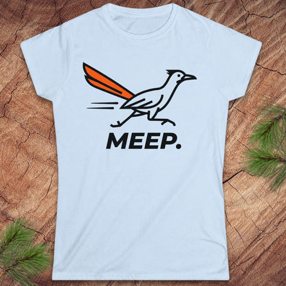 A light blue short-sleeve crewneck t-shirt featuring a graphic of a roadrunner with an orange tail and the word 'MEEP.' printed below in black, laid flat on a rustic wood background with pine branches in the corners.