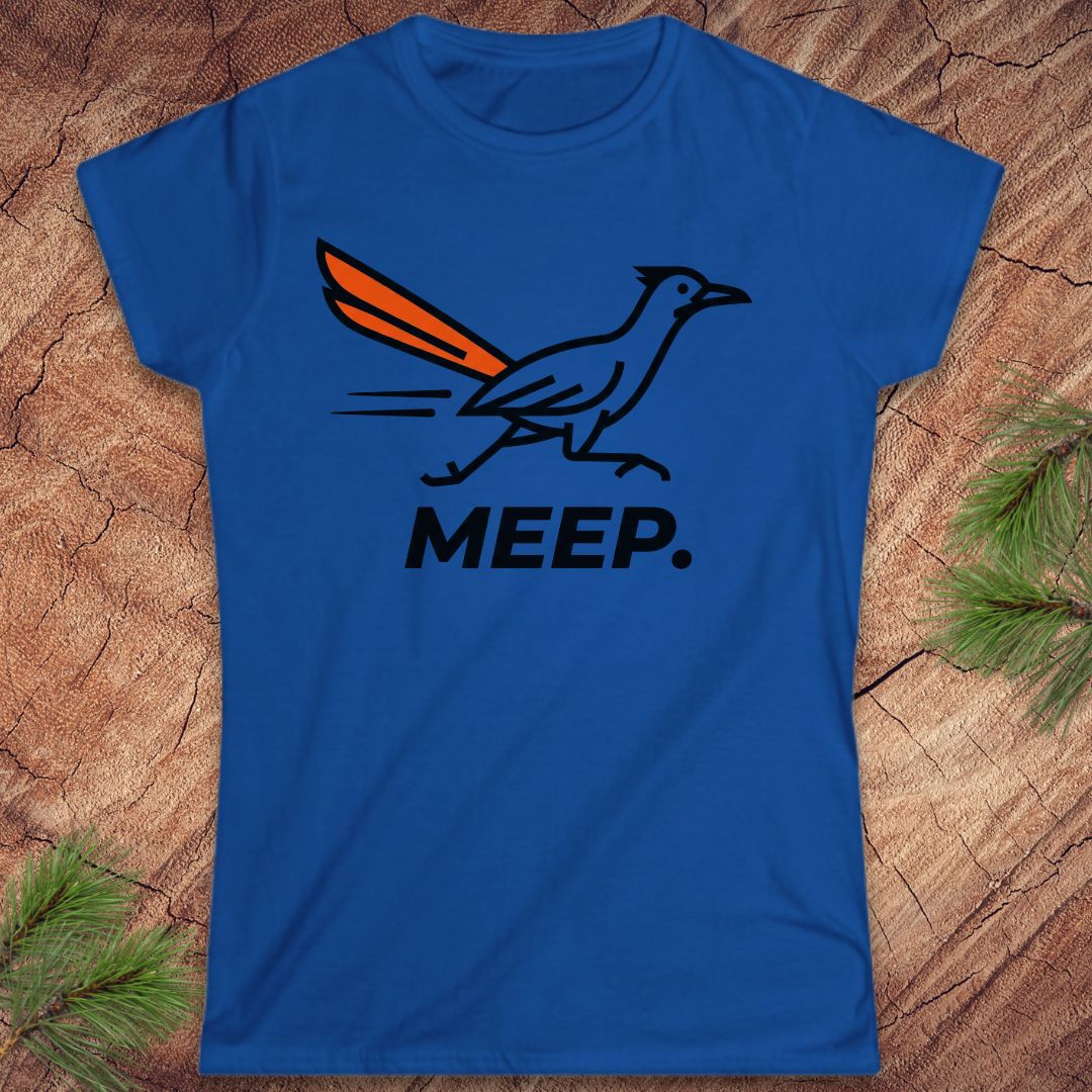 A royal blue short-sleeve t-shirt with a graphic of a roadrunner and the word 'MEEP.' printed on the front, displayed flat on a rustic wood background with pine needles.
