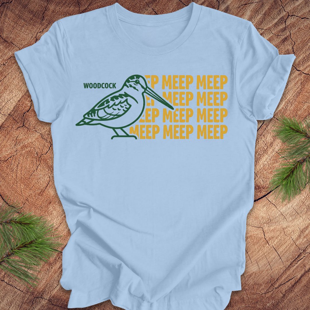 Light blue t-shirt with a green woodcock graphic and yellow 'meep' text on a wooden surface.