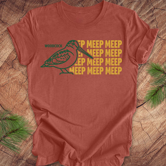 Clay colored t-shirt with a green woodcock graphic and yellow 'meep' text on a wooden surface.