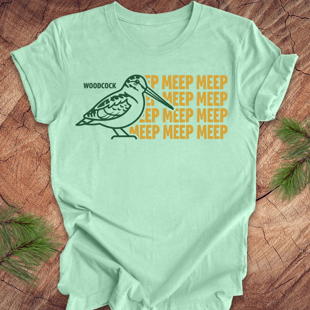 Mint green t-shirt with a green woodcock graphic and yellow 'meep' text on a wooden surface.