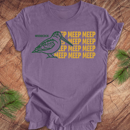 Purple t-shirt with a green woodcock graphic and yellow 'meep' text on a wooden surface.