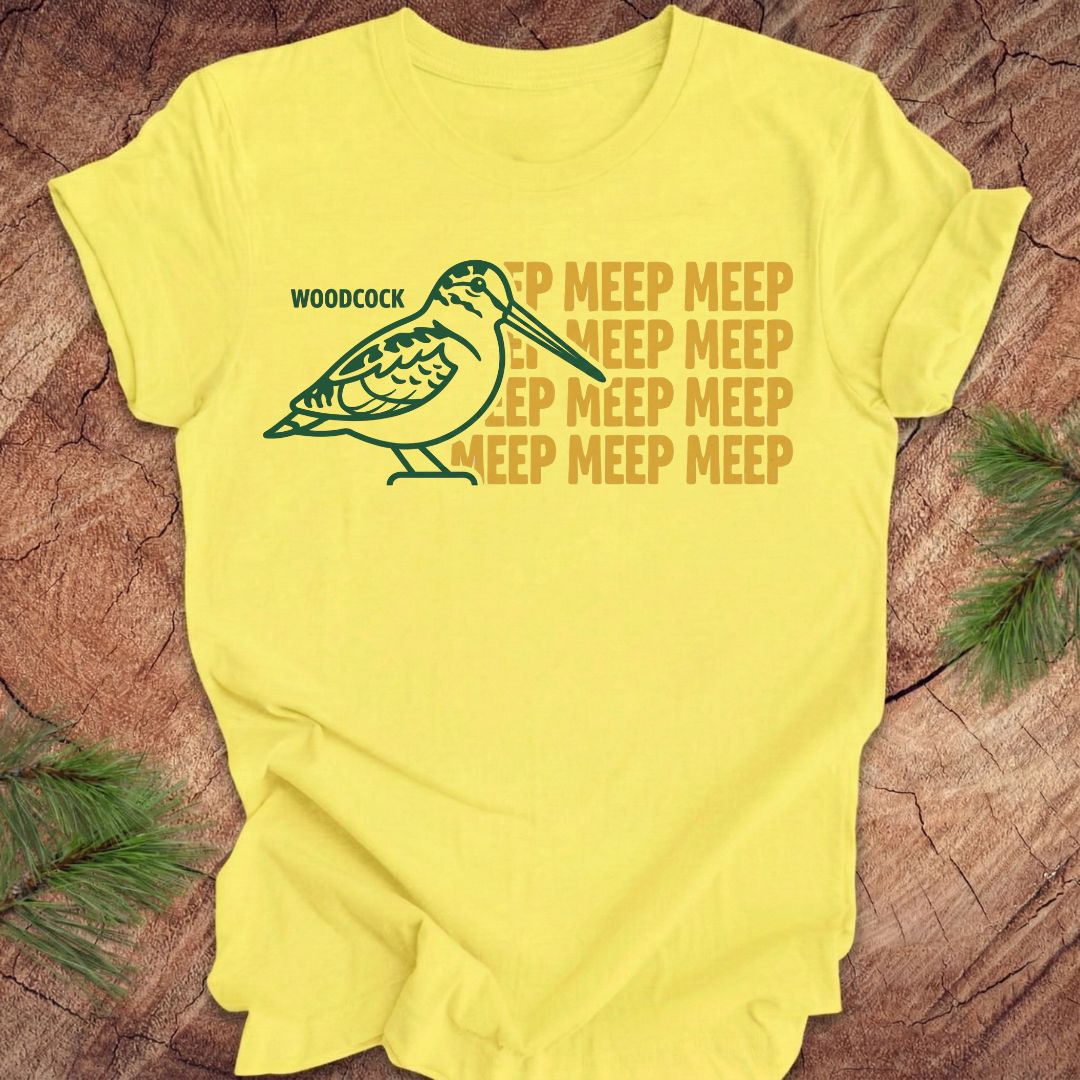 Yellow t-shirt with a green woodcock graphic and yellow 'meep' text on a wooden surface.
