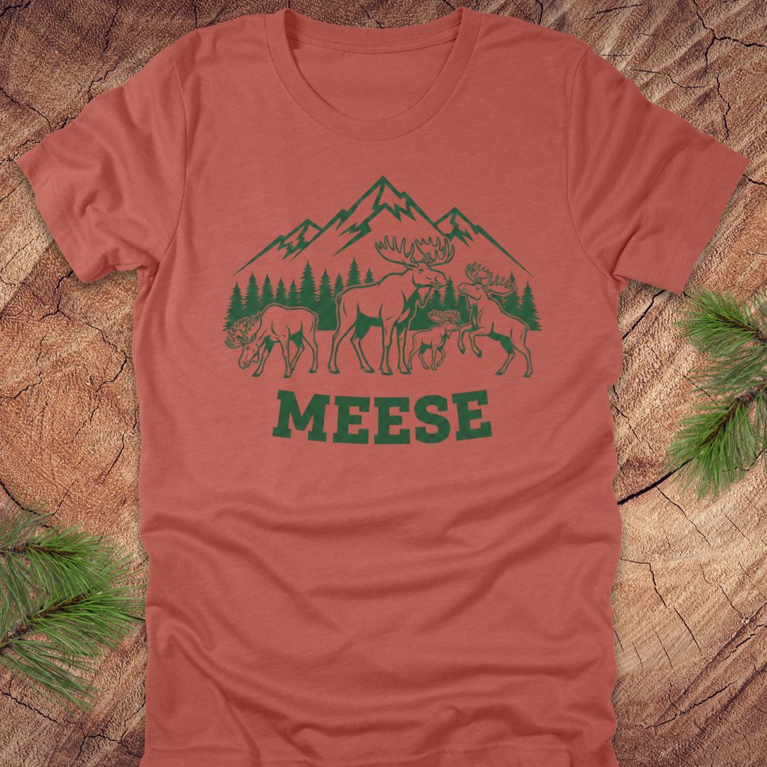 Clay colored t-shirt with moose, trees and mountains with text "meese"on a wood background. 