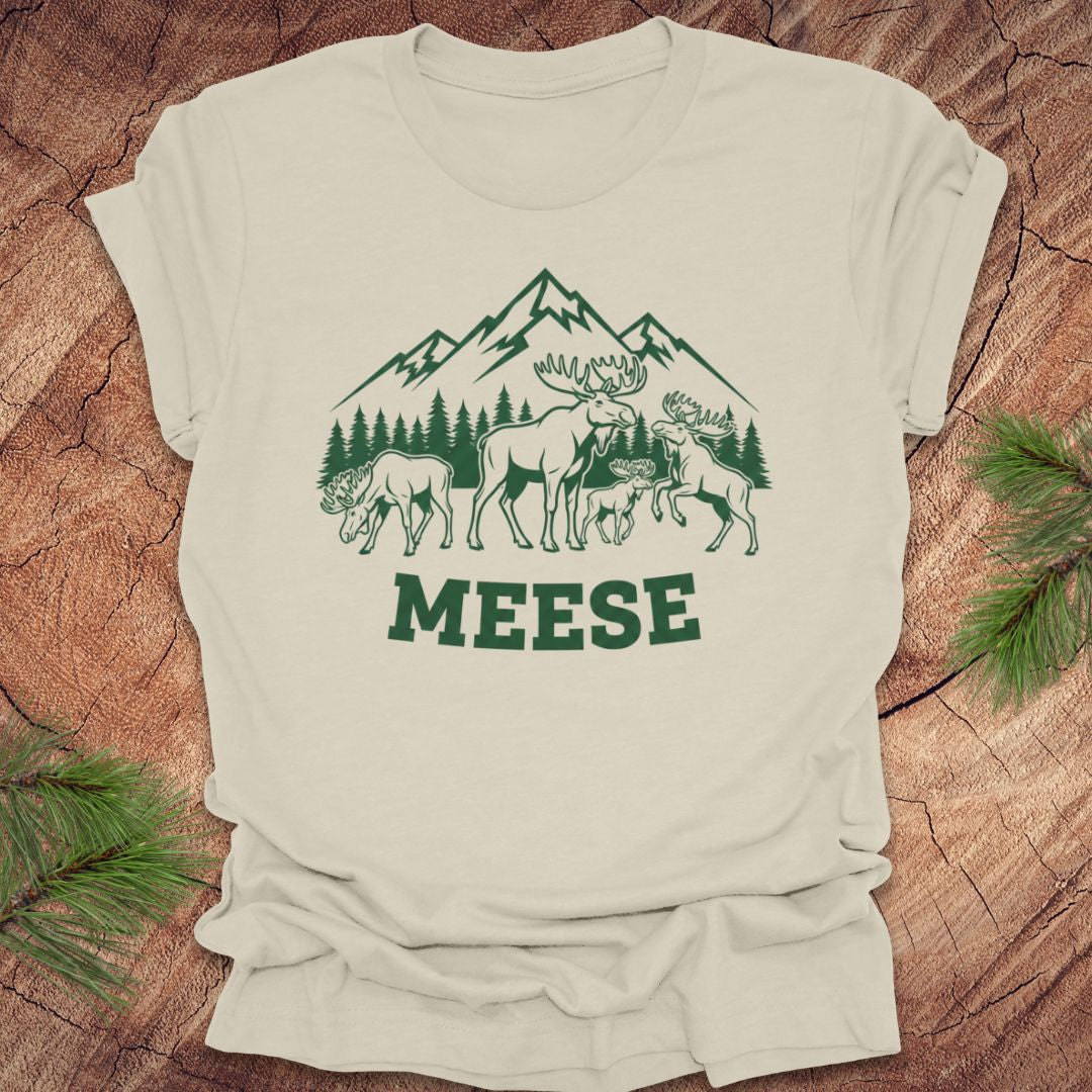 Beige t-shirt with moose, trees and mountains with text "meese"on a wood background. 