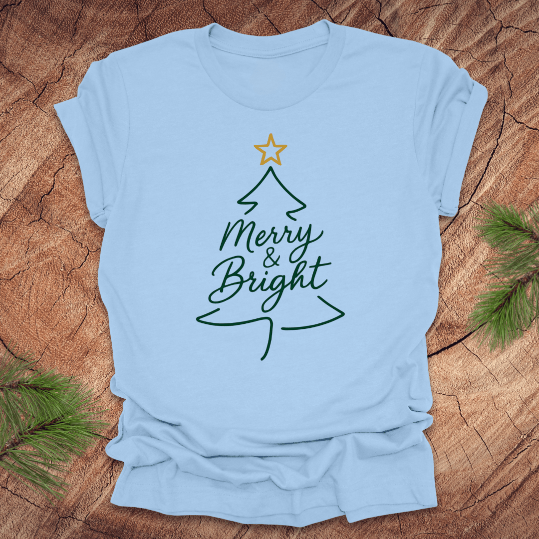 Light blue t-shirt with Christmas tree design and 'Merry & Bright' text on a wooden surface.