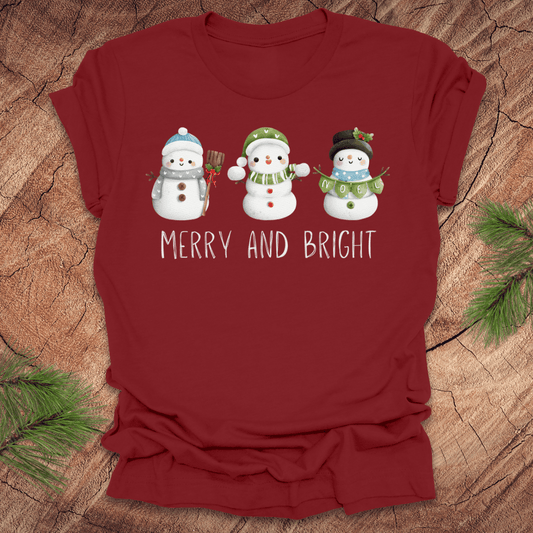 Dark Red t-shirt with snowman design and 'Merry and Bright' text on a wooden surface.