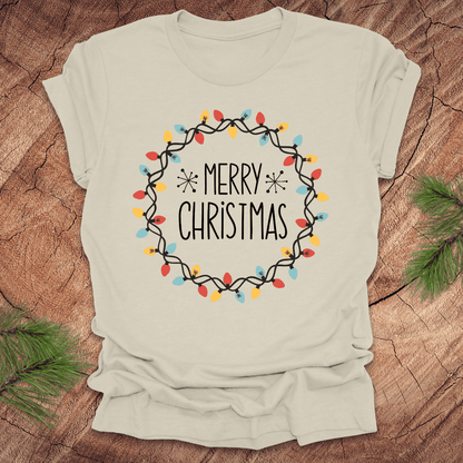 Beige t-shirt with 'Merry Christmas' design on a wooden surface with pine branches.
