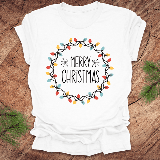 White t-shirt with 'Merry Christmas' design on a wooden surface with pine branches.