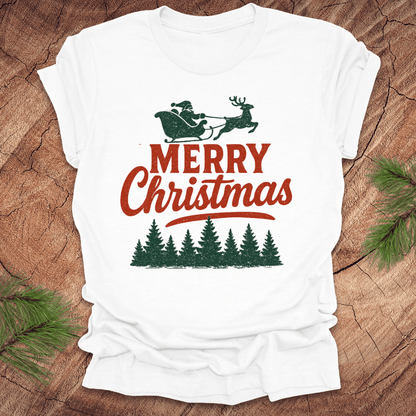 White t-shirt with 'Merry Christmas' design on a wooden surface with pine branches.
