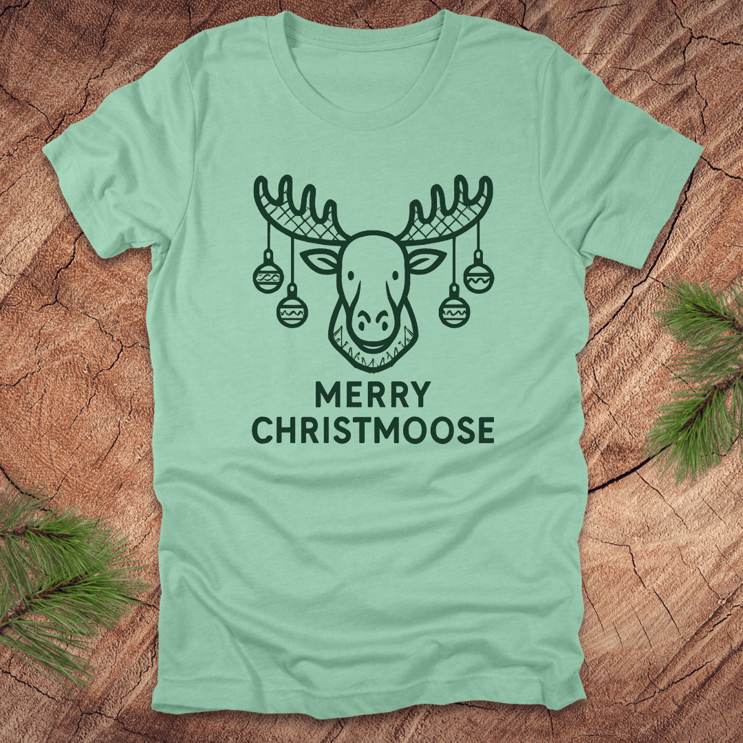 Light green t-shirt with a moose graphic and 'Merry Christmoose' text on a wooden surface.