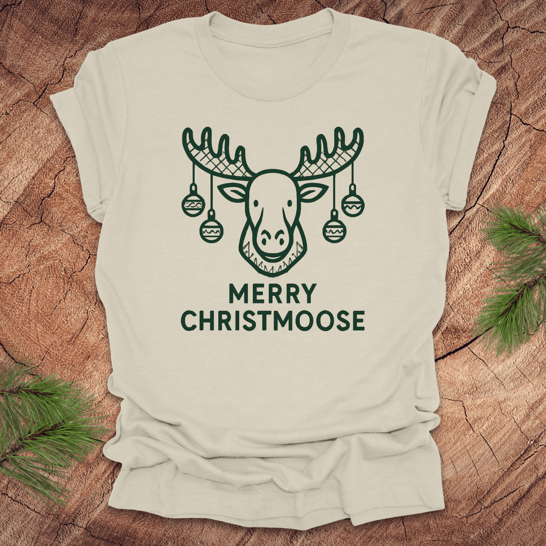 Beige t-shirt with a moose graphic and 'Merry Christmoose' text on a wooden surface.