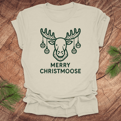 Beige t-shirt with a moose graphic and 'Merry Christmoose' text on a wooden surface.