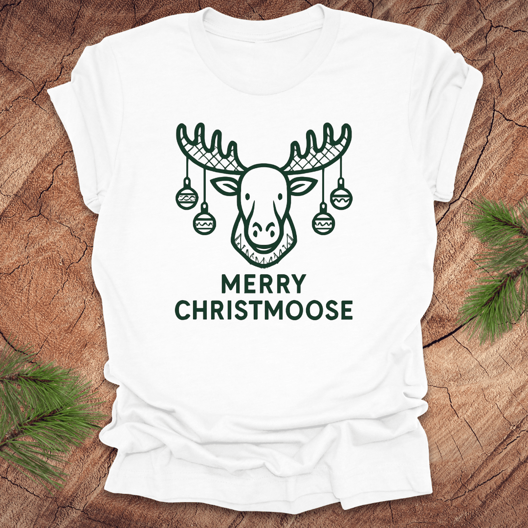White t-shirt with 'Merry Christmoose' design on a wooden surface with pine branches.