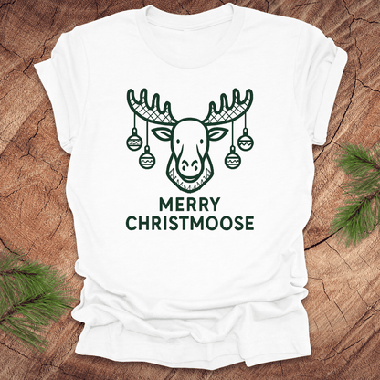 White t-shirt with 'Merry Christmoose' design on a wooden surface with pine branches.