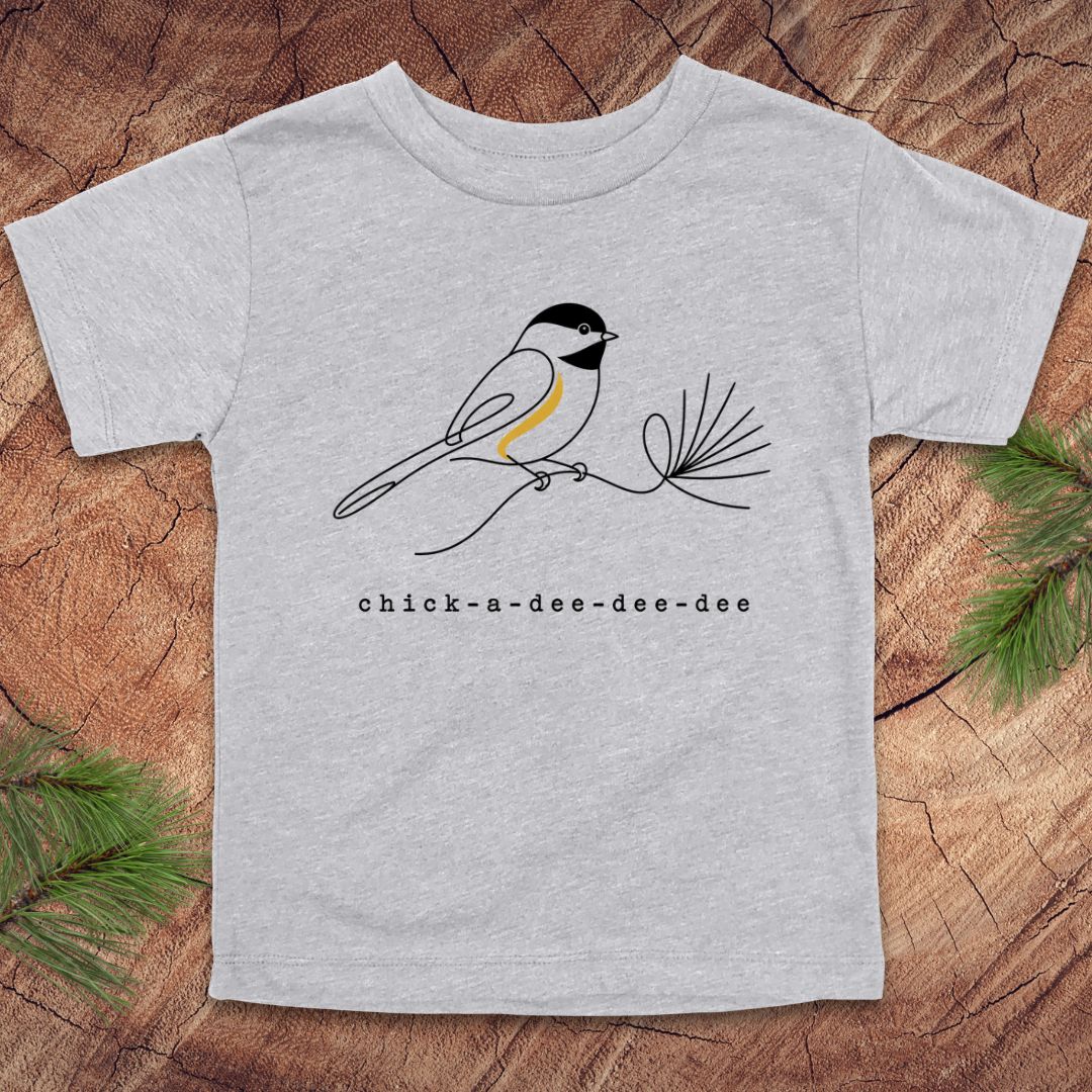 A light heather gray short-sleeve t-shirt featuring a black and white chick-a-dee bird with a yellow chest accent perched on a pine branch, with the text 'chick-a-dee-dee-dee' printed below. The t-shirt is laid flat on a rustic wood background with pine needles in the corners.
