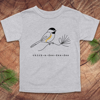 A light heather gray short-sleeve t-shirt featuring a black and white chick-a-dee bird with a yellow chest accent perched on a pine branch, with the text 'chick-a-dee-dee-dee' printed below. The t-shirt is laid flat on a rustic wood background with pine needles in the corners.