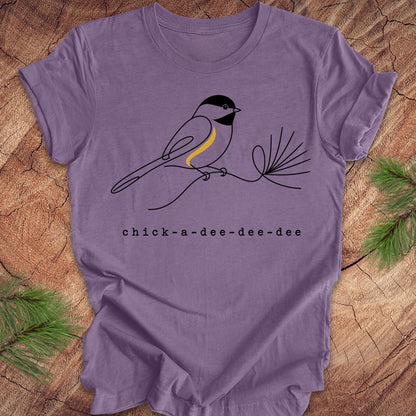 A purple short-sleeve t-shirt laid flat on a rustic wood background with pine branches in the corners. The shirt features a minimalist black line drawing of a chickadee with a yellow chest patch and the text 'chick-a-dee-dee-dee' printed below.