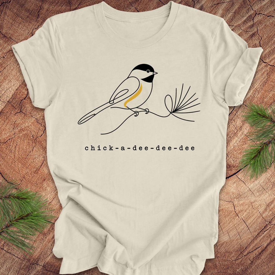 Beige t-shirt with chickadee design and 'chick-a-dee-dee-dee' text on a wooden surface with pine branches.