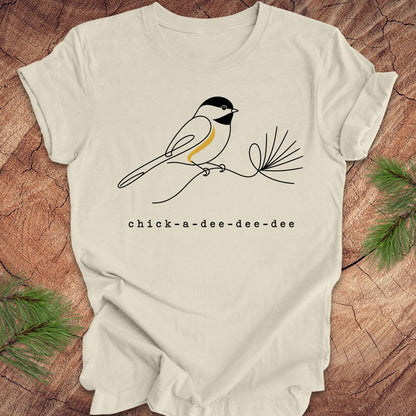 Beige t-shirt with chickadee design and 'chick-a-dee-dee-dee' text on a wooden surface with pine branches.