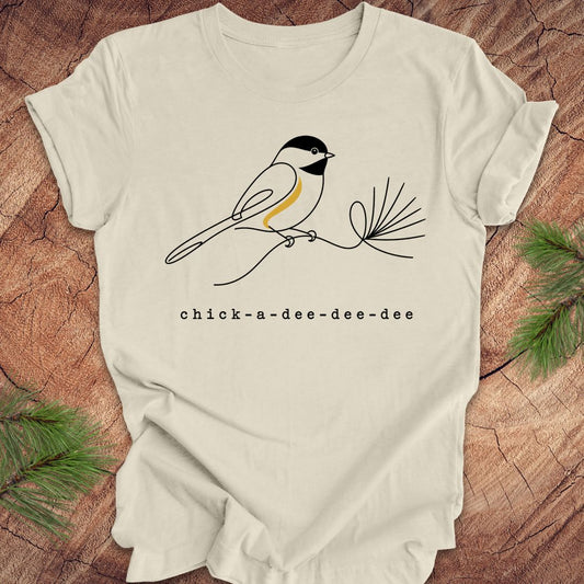 Beige t-shirt with chickadee design and 'chick-a-dee-dee-dee' text on a wooden surface with pine branches.