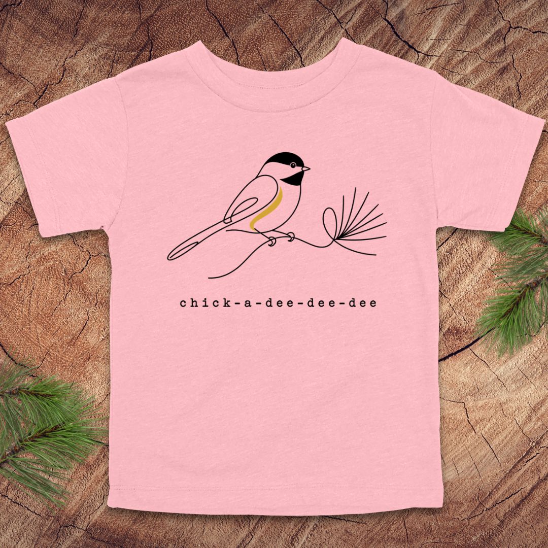 A light pink short-sleeve t-shirt featuring a minimalist black line art illustration of a chickadee perched on a pine branch, with the text 'chick-a-dee-dee-dee' printed below. The t-shirt is laid flat on a rustic wood slice background decorated with green pine needles.