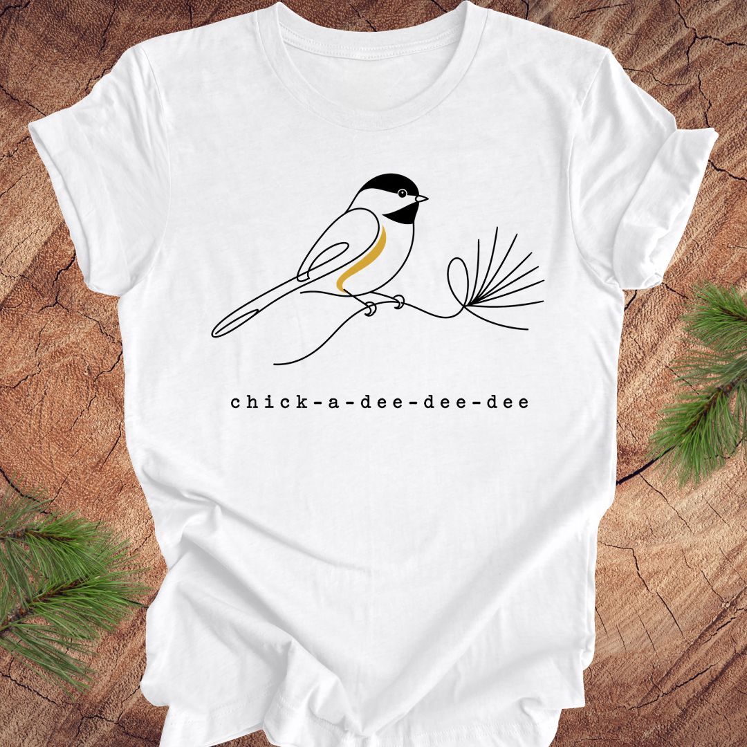 White t-shirt with a chickadee design and text on a wooden surface with greenery.