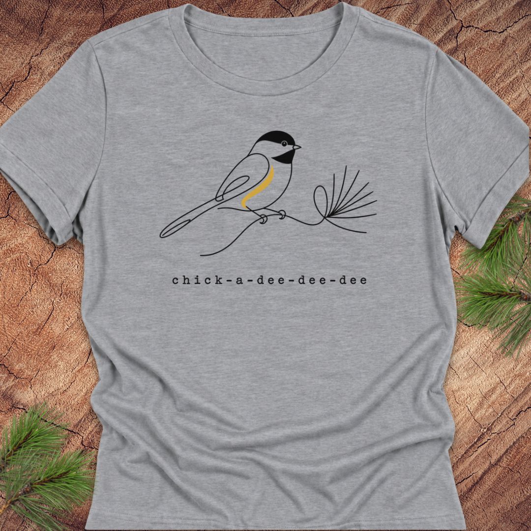 Athletic heather gray women's relaxed t-shirt with a one-line Black-capped Chickadee on a pine sprig above 'chick-a-dee-dee-dee' text, laid flat on a rustic wood background with pine branches.