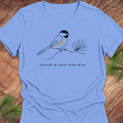 Carolina blue women's relaxed t-shirt with a one-line Black-capped Chickadee on a pine sprig above 'chick-a-dee-dee-dee' text, laid flat on a rustic wood background with pine branches.