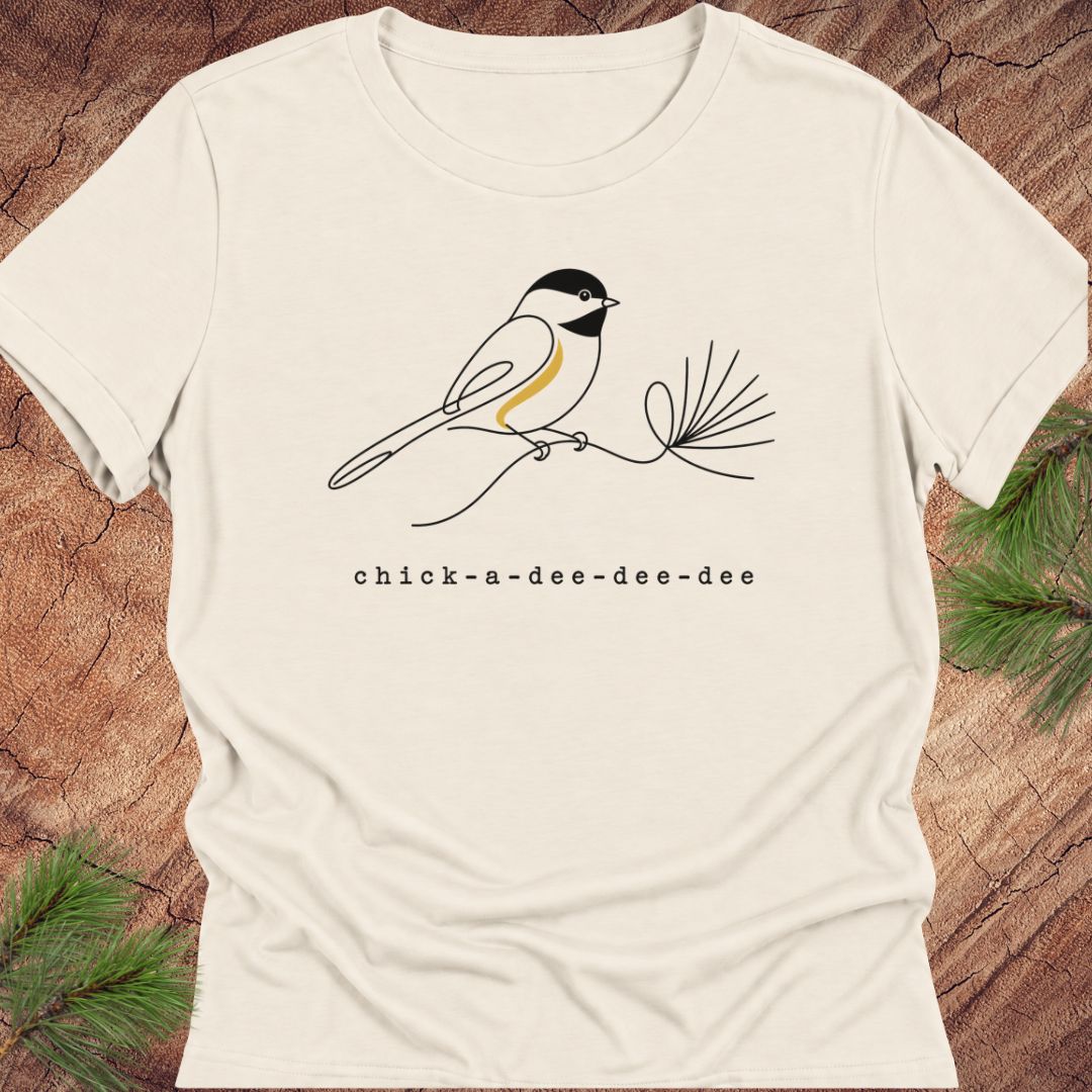 Natural cream women's relaxed t-shirt with a one-line Black-capped Chickadee on a pine sprig above 'chick-a-dee-dee-dee' text, laid flat on a rustic wood background with pine branches.