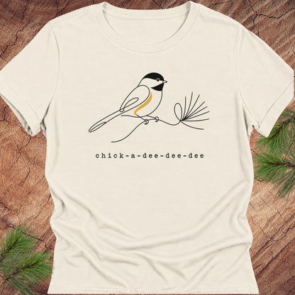 Natural cream women's relaxed t-shirt with a one-line Black-capped Chickadee on a pine sprig above 'chick-a-dee-dee-dee' text, laid flat on a rustic wood background with pine branches.