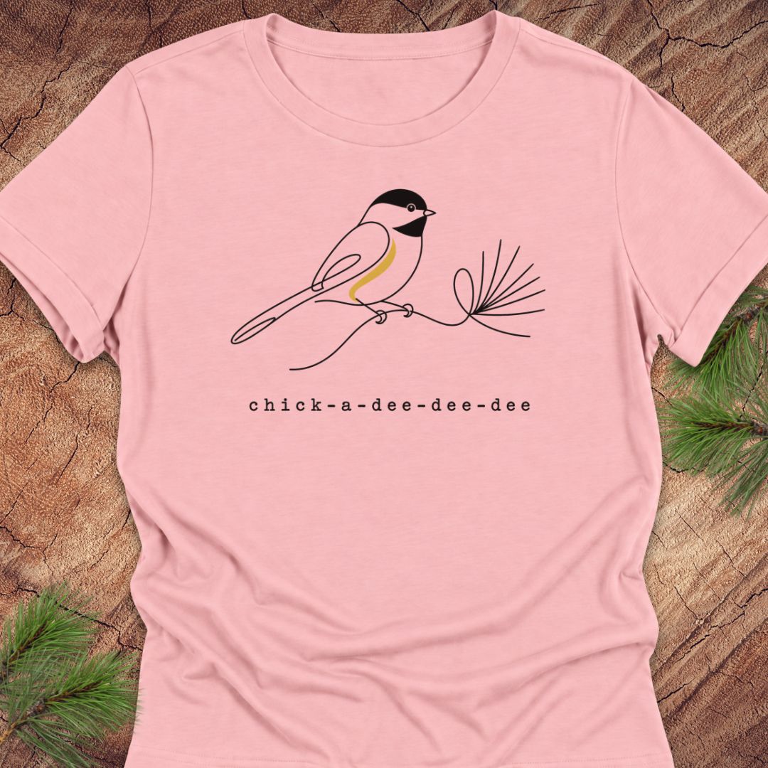 Pink women's relaxed t-shirt with a one-line Black-capped Chickadee on a pine sprig above 'chick-a-dee-dee-dee' text, laid flat on a rustic wood background with pine branches.