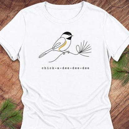 White women's relaxed t-shirt with a one-line Black-capped Chickadee on a pine sprig above 'chick-a-dee-dee-dee' text, laid flat on a rustic wood background with pine branches.
