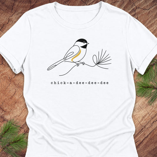 White women's relaxed t-shirt with a one-line Black-capped Chickadee on a pine sprig above 'chick-a-dee-dee-dee' text, laid flat on a rustic wood background with pine branches.