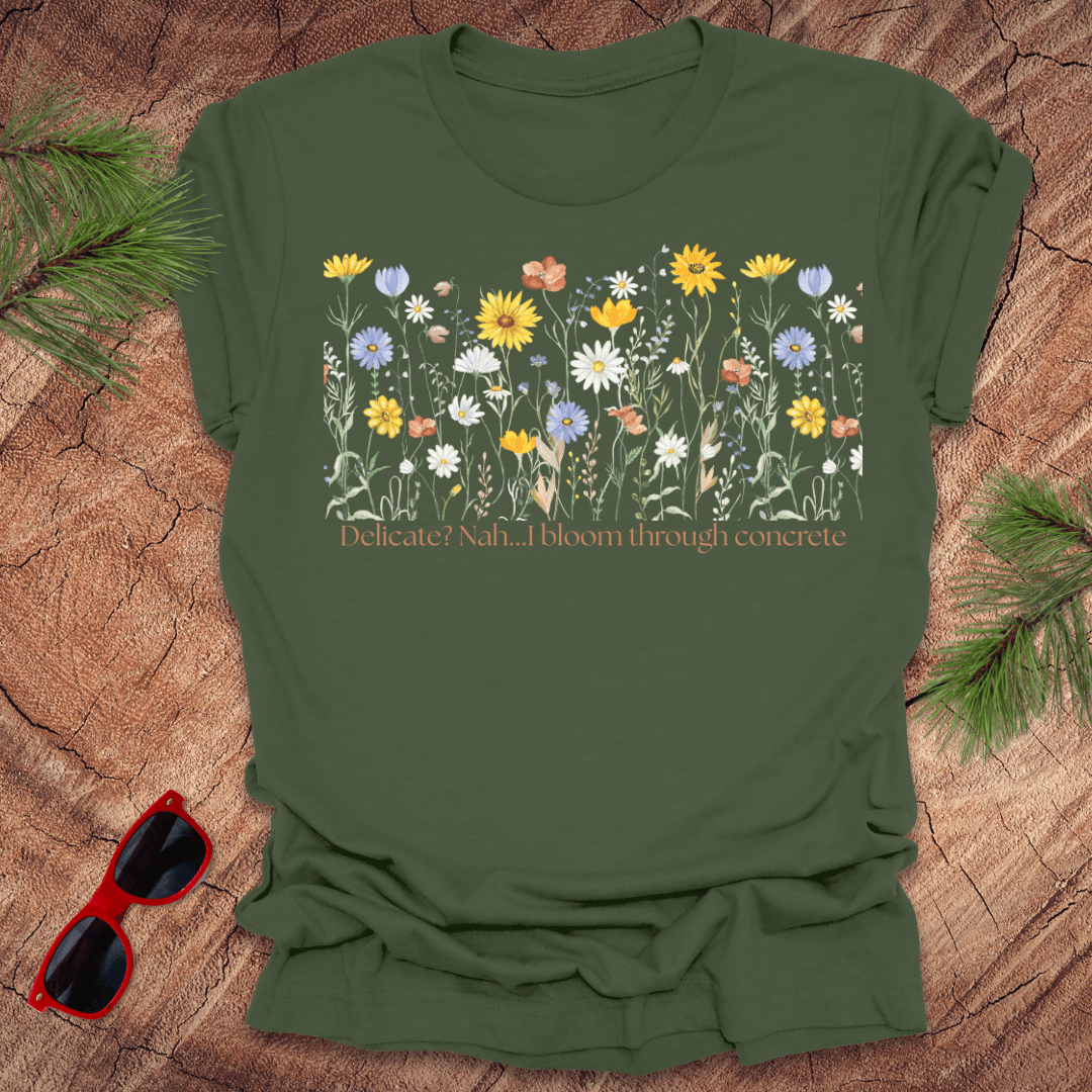Green t-shirt with floral design and text "delicate? nah. i bloom through concrete.' on a wooden surface.